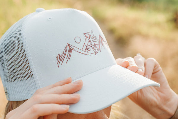 Hands holding a white mesh-back hat with a crisp embroidered mountain design on the front panel.
