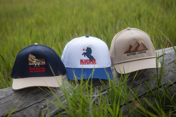 Three embroidered hats side by side on a log in tall grass, each with a different crown and brim.