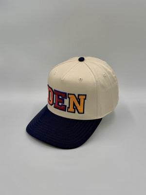 DEN 5-panel snapback seen from an angle, natural crown and navy brim, embroidered by Threaded by Sal